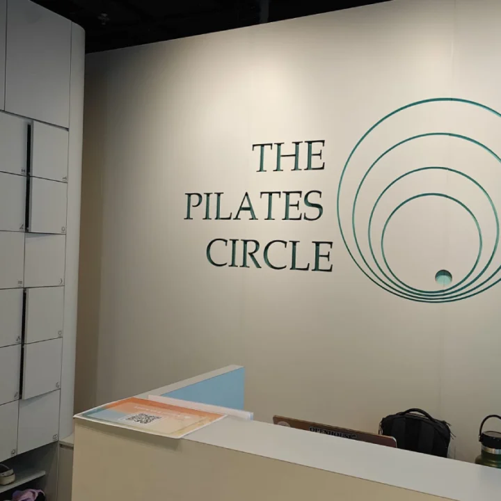 Reception area of The Pilates Circle with the studio's circular logo and wordmark on a white wall, alongside a wall-mounted cubby storage unit and a front desk counter.