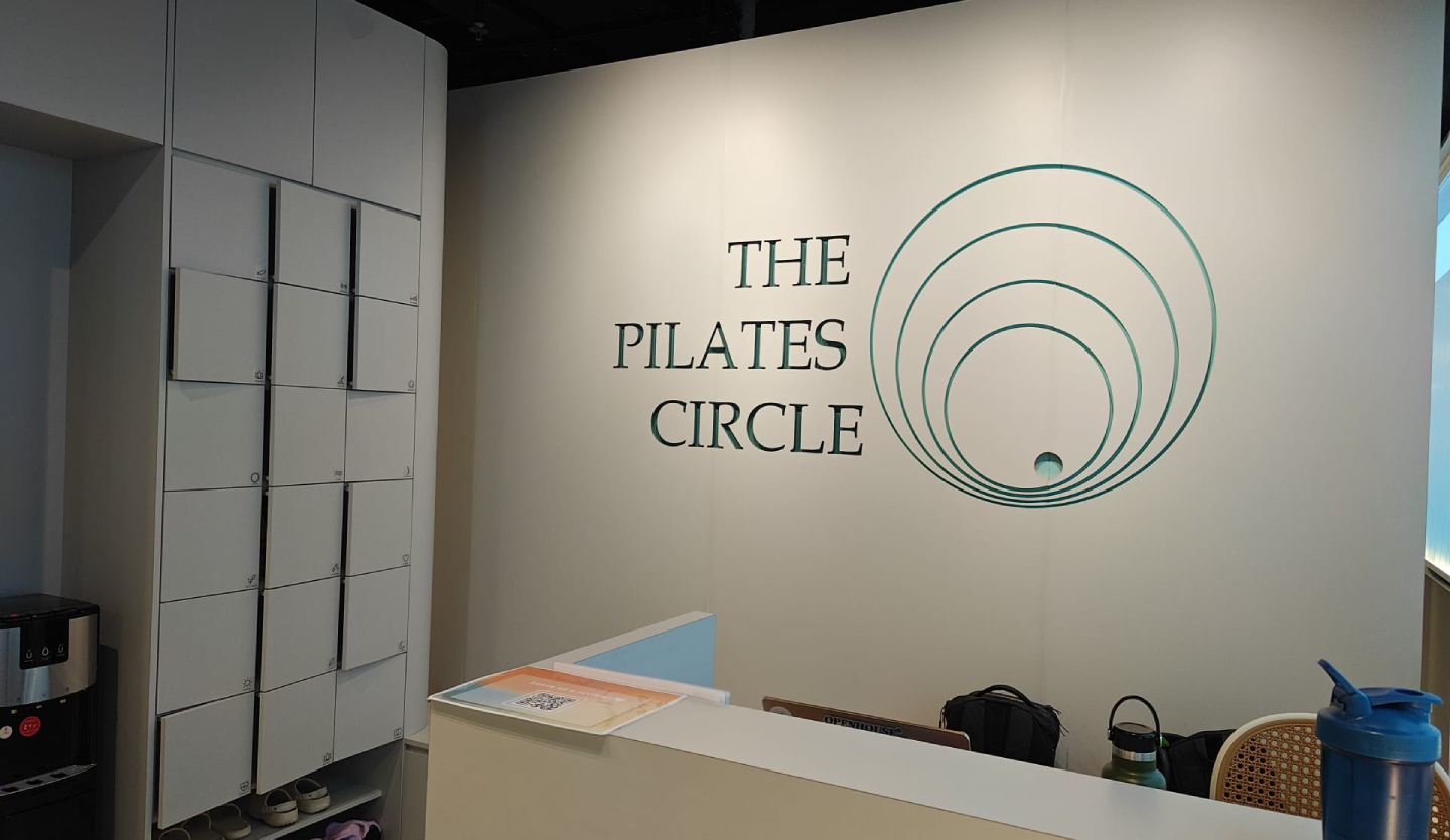 Reception area of The Pilates Circle with the studio's circular logo and wordmark on a white wall, alongside a wall-mounted cubby storage unit and a front desk counter.