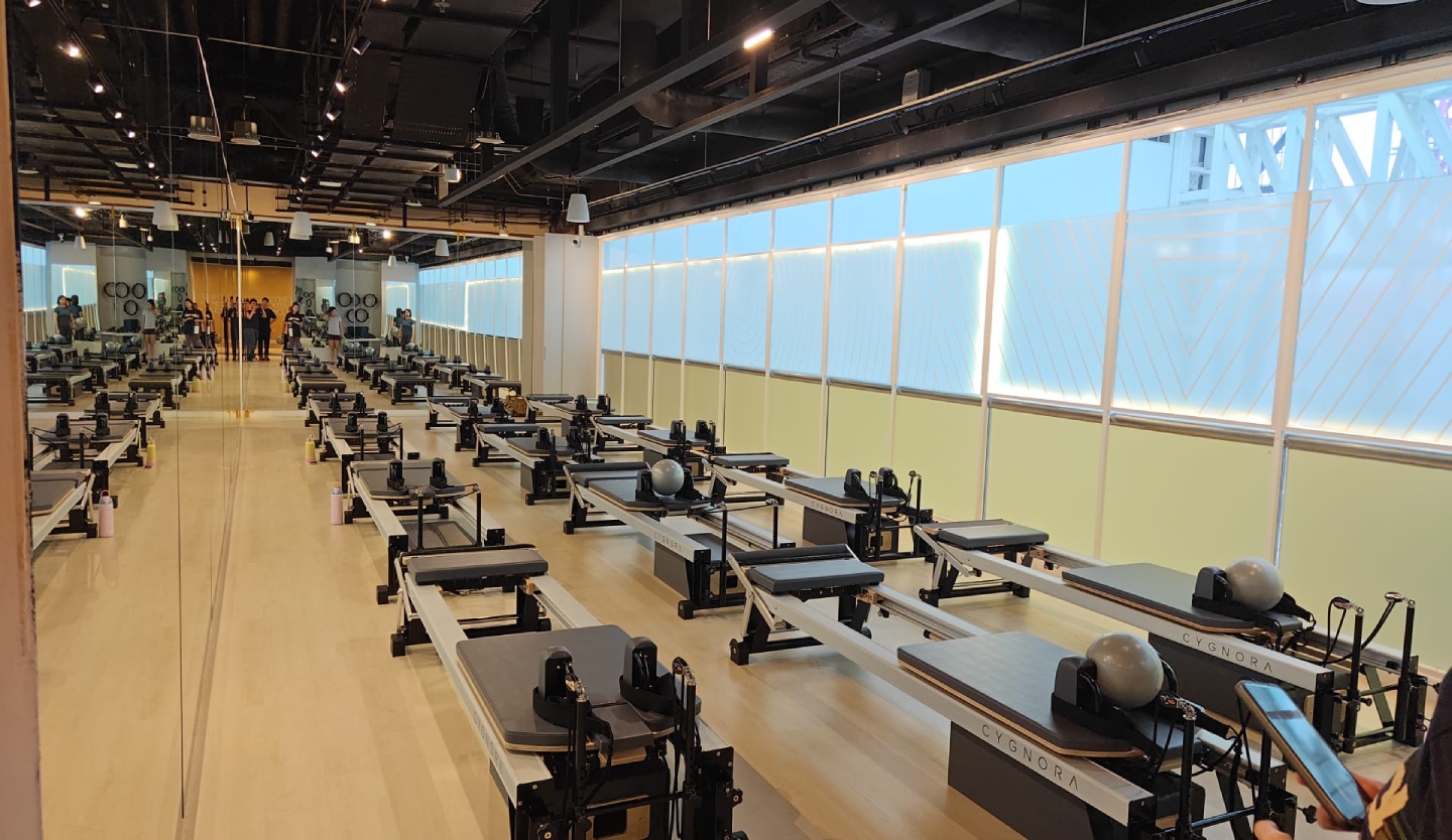 Interior of The Pilates Circle studio showing rows of Cygnora Pilates reformer machines facing floor-to-ceiling windows, with a mirrored wall reflecting the space on the left.